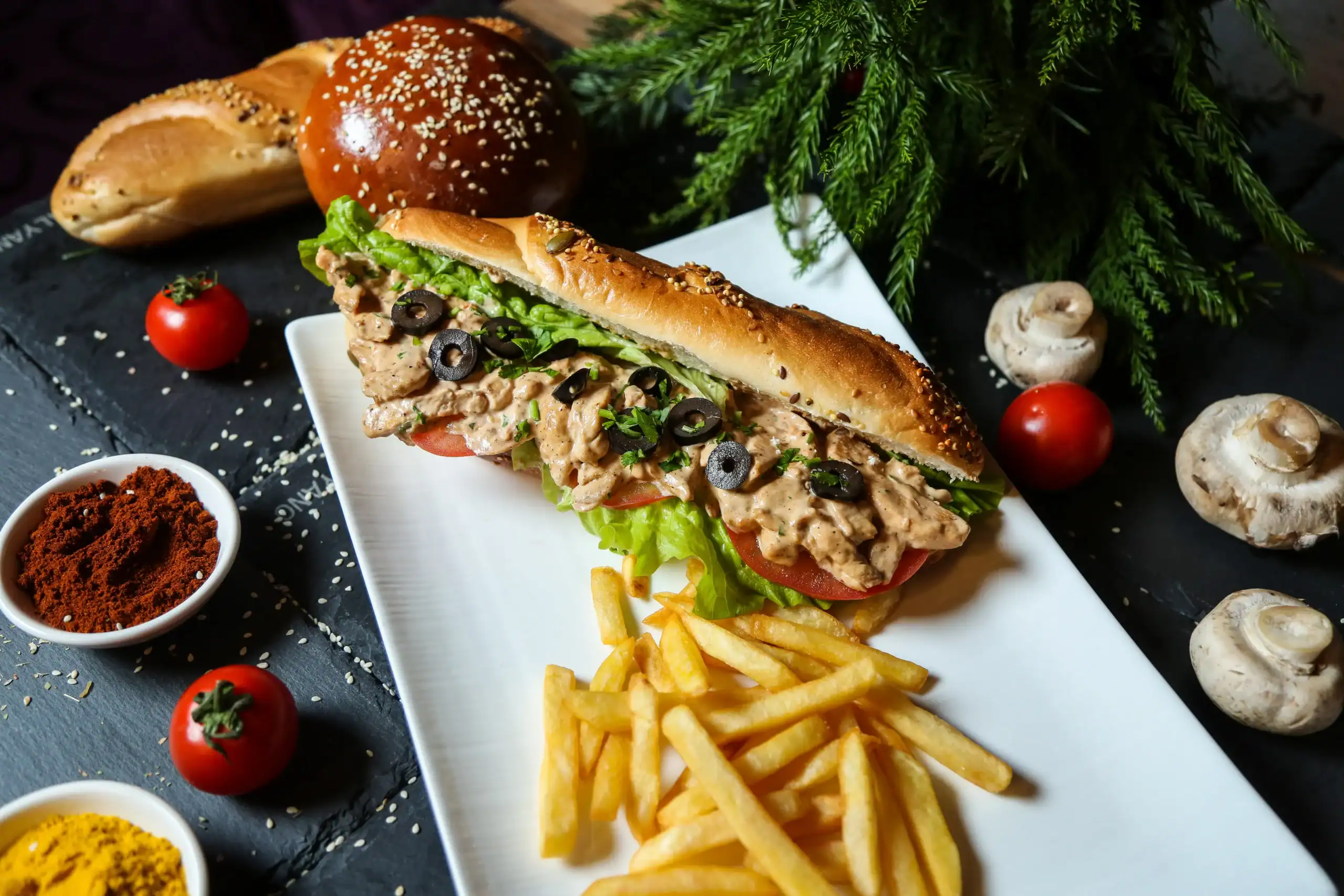 Savory tuna sandwich with fresh vegetables on a baguette, crispy fries on white plate, surrounded by cherry tomatoes, mushrooms, and spices, healthy meal, delicious street food, RecipeStang.