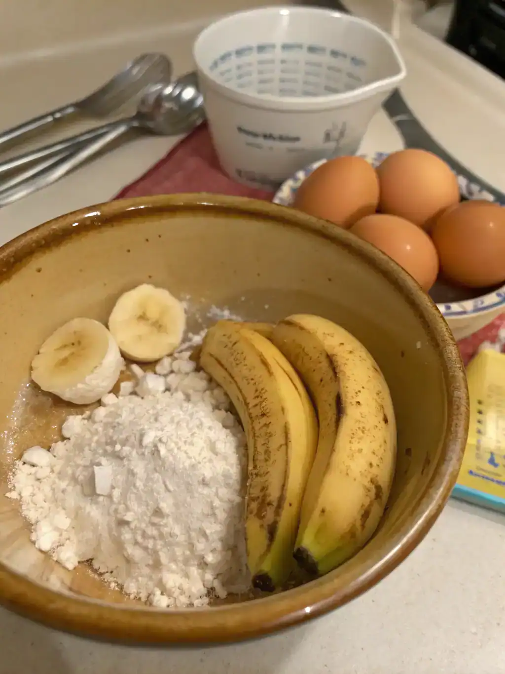 Sliced bananas, flour, and eggs in a rustic bowl for baking or cooking. Fresh ingredients ready for a recipe, with other kitchen items like eggs and measuring cups in the background. Perfect for breakfast or dessert preparations.