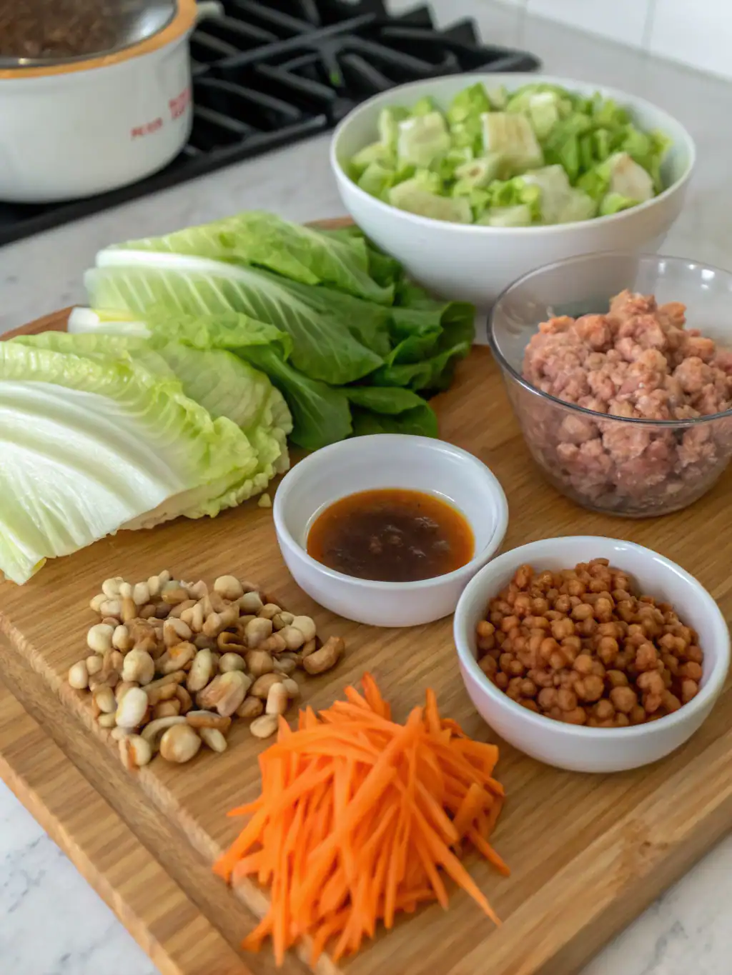 Fresh vegetable and bean ingredients for Asian-inspired cooking on a wooden cutting board. Includes lettuce, shredded carrots, chickpeas, lentils, chopped celery, ground meat, and dipping sauce.