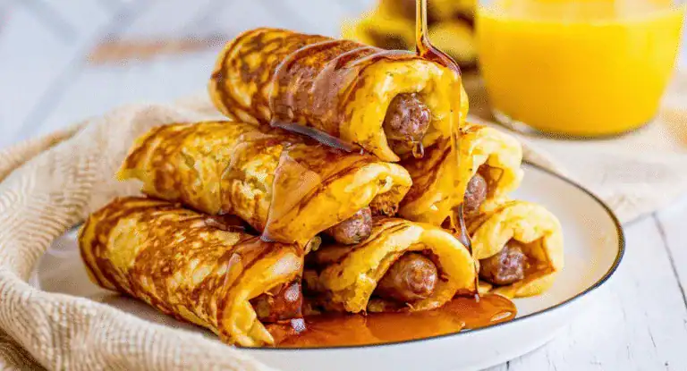 Crispy breakfast crepes filled with sausage, drizzled with syrup, served on a white plate with a side of orange juice, perfect for morning meals.