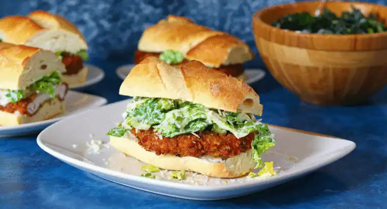 Crispy fried chicken sandwich with fresh lettuce and creamy sauce on a baguette, served on a white plate with additional sandwiches and a bowl of greens in the background.
