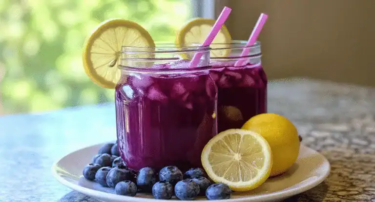 Vibrant purple blueberry lemonade in glass jars with lemon slices and pink straws, served on a white plate with fresh blueberries and whole lemons, perfect for refreshing summer drinks.