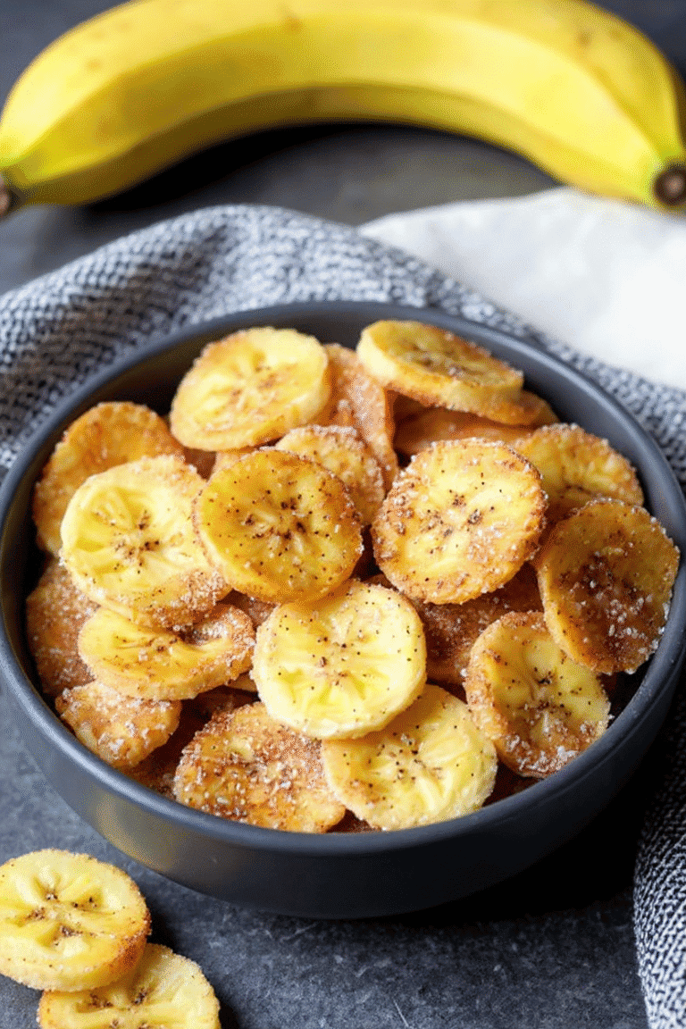 Banana Chips in a black bowl on gray surface, fresh banana in background, crispy and sugared banana snack, ideal for healthy snacking or dessert, homemade banana chips recipe, close-up.
