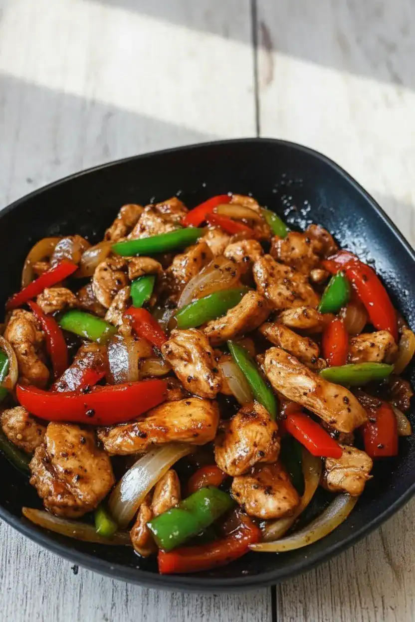 Stir-fried chicken with colorful bell peppers and onions in a black bowl, showcasing a flavorful Asian-inspired dish.